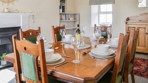 A dining room with a wooden table set for six, surrounded by chairs. There is a fireplace, a bookshelf, and a window with curtains.