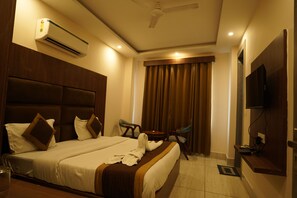 A hotel room with a large bed, a ceiling fan, an air conditioning unit, a TV mounted on the wall, and a small table with a chair. - Hotel Divine Aura - A Premium Hotel (Haridwar)