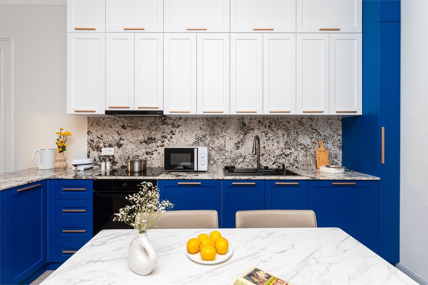 Una cocina moderna con armarios superiores blancos, bajos azules, encimera de granito, microondas, pileta y una mesa de comedor con un florero y limones.