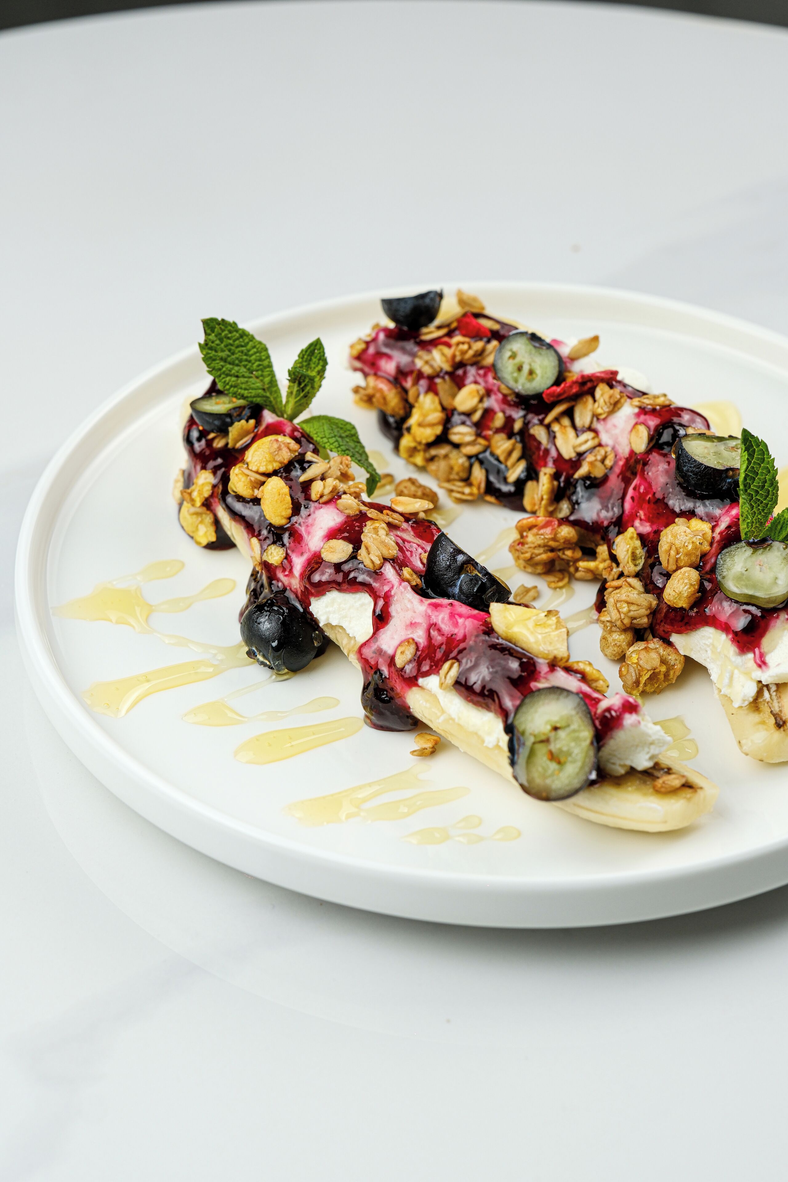 A white plate with a dessert consisting of fruit, granola, and a drizzle of syrup.