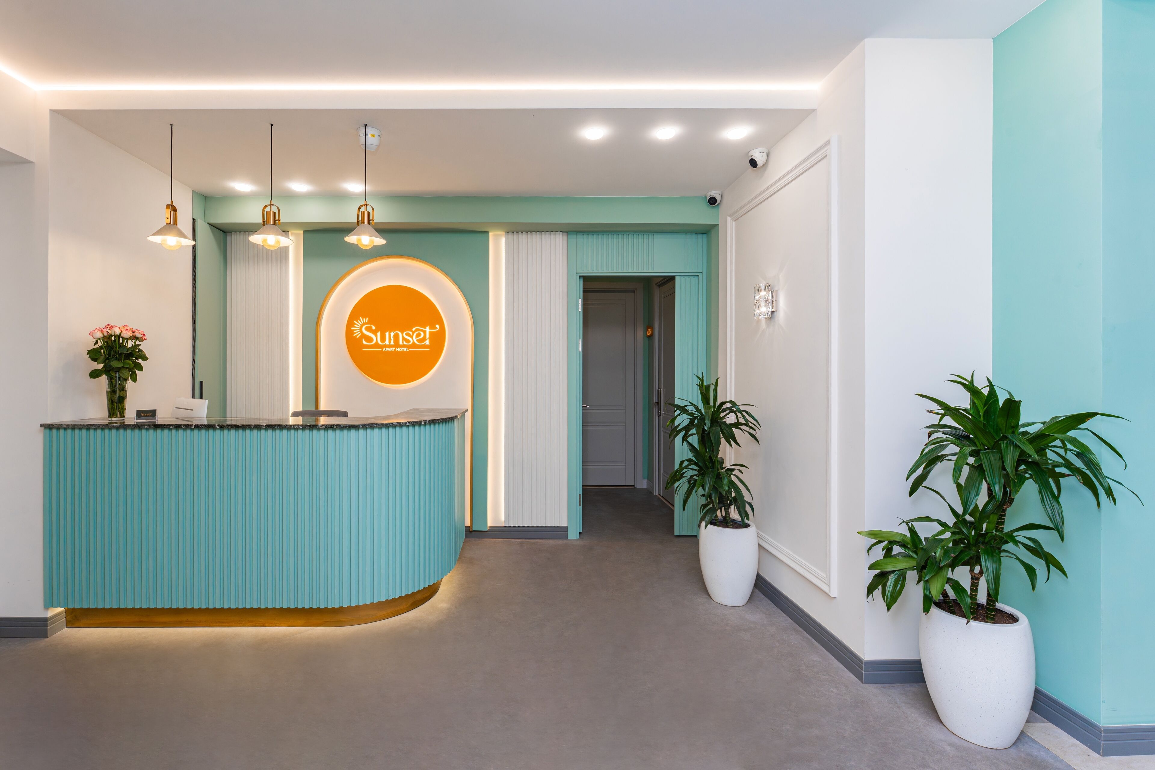 Een receptiegedeelte met een receptiebalie in teal, een rond bord met het 'Sunrise'-logo, hanglampen en potplanten.