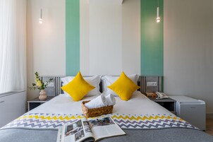 Een net opgemaakt bed met een bedsprei met patroon, twee heldergele kussens, een rieten mand met handdoeken en een nachtkastje met een telefoon en een boek.