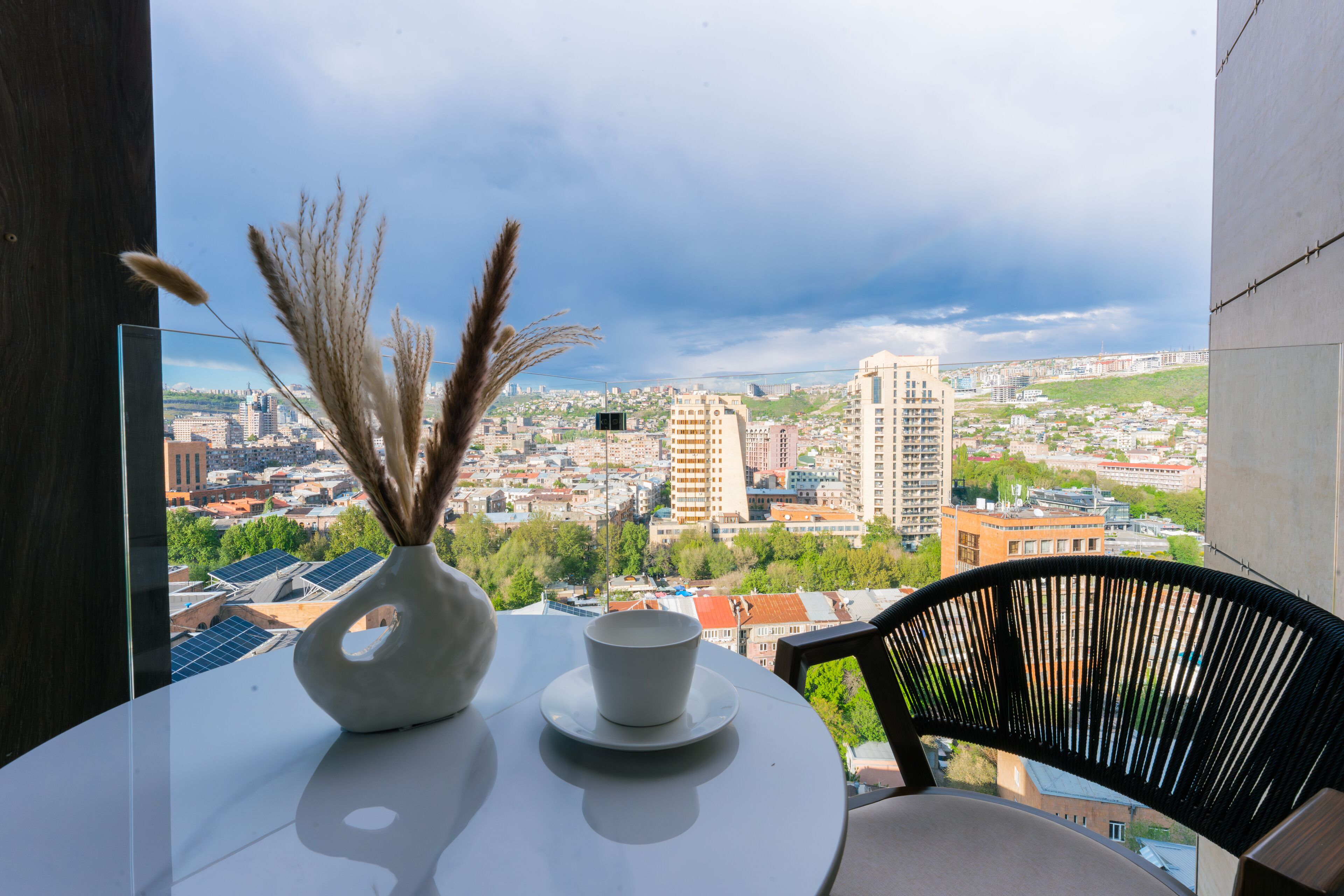A balcony with a table, a chair, a vase with dried plants, and a cup. The view includes a cityscape with buildings and greenery.