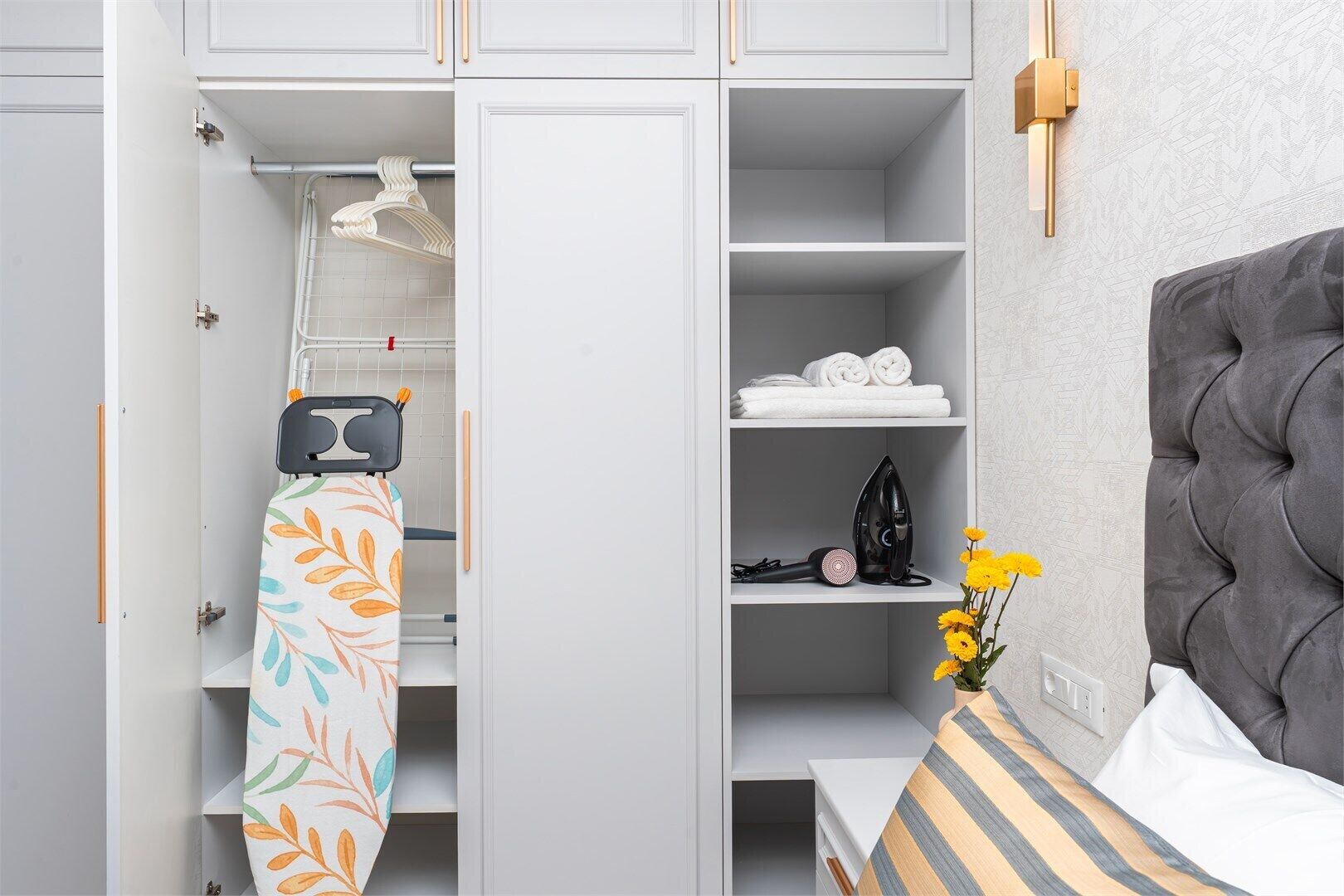 A bedroom with a grey tufted headboard, a bedside table with a striped pillow, a small vase with yellow flowers, and a wardrobe with shelves containing folded towels and an iron.