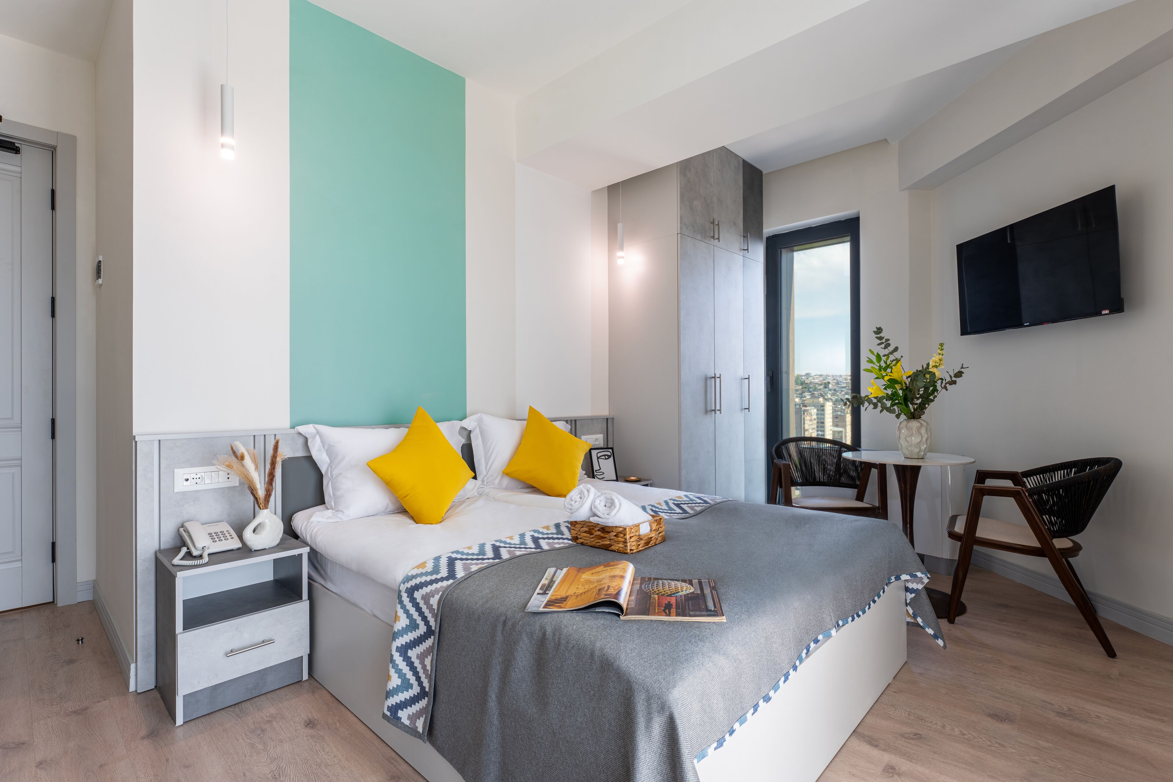 A modern hotel room with a bed, bedside table, nightstand, and a small seating area with a chair and table.