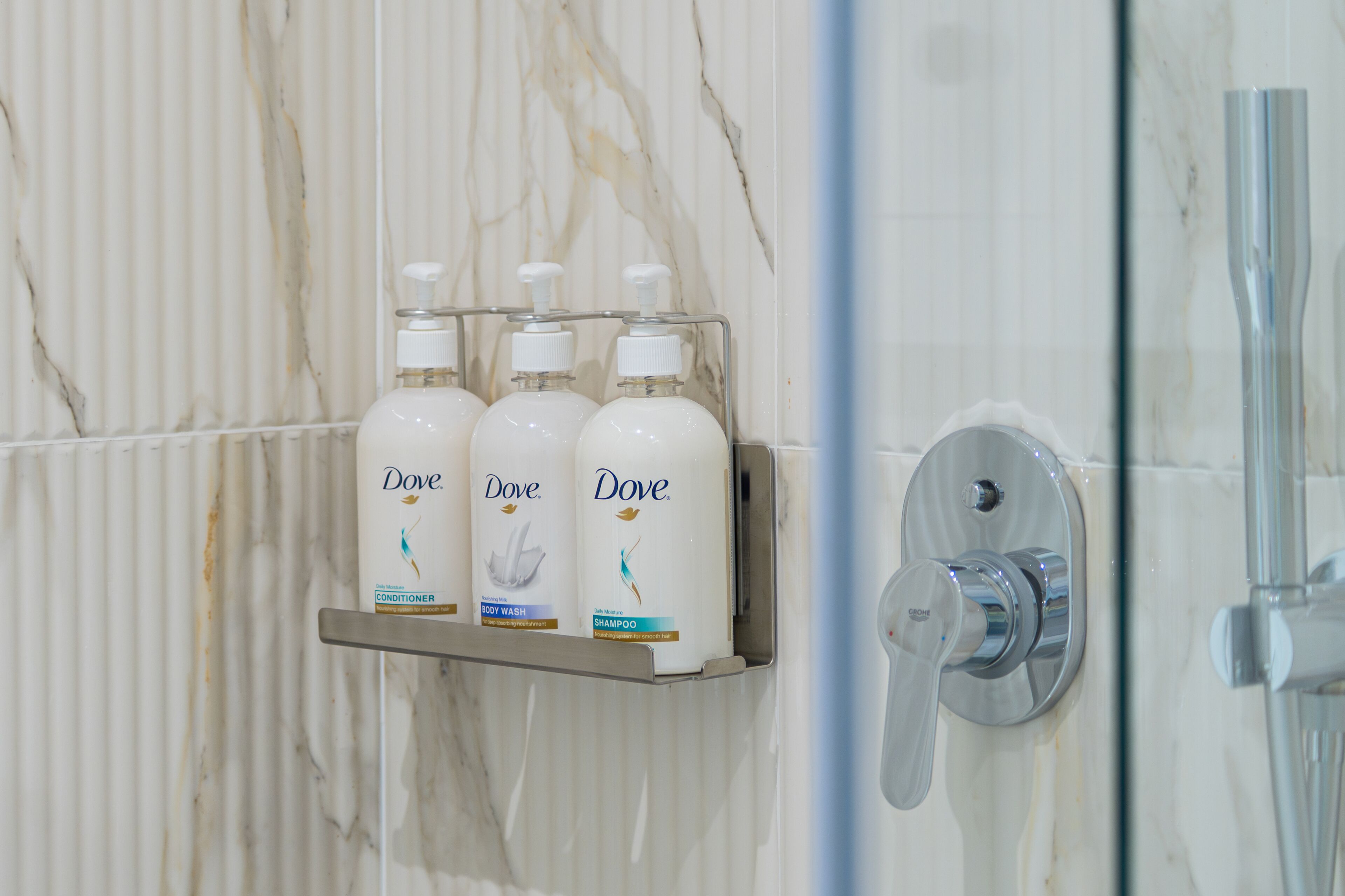 Une salle de bain avec un mur en marbre, une douche avec des accessoires chromés et une étagère où sont posés des flacons de gel douche et de shampoing Dove.