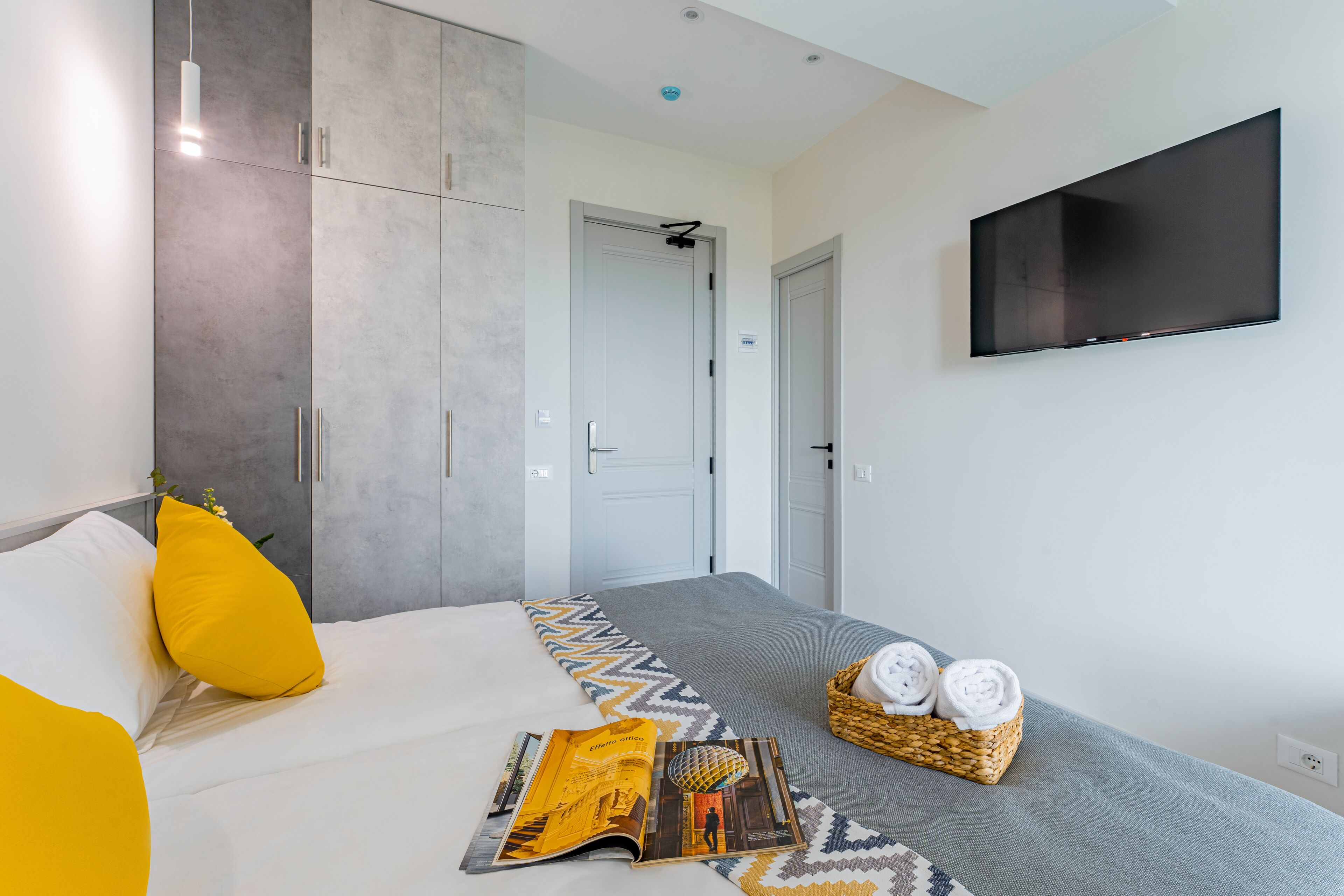 A modern hotel room with a bed, a flat-screen TV, a magazine, and a basket of towels.