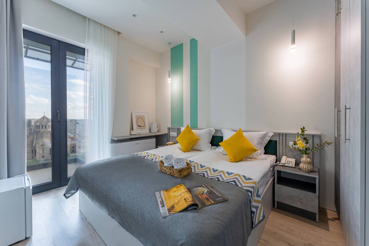 Un hotel moderno con una cama grande, una mesita de noche con lámpara, vistas de la ciudad a través de un ventanal, y un paisaje urbano al exterior.
