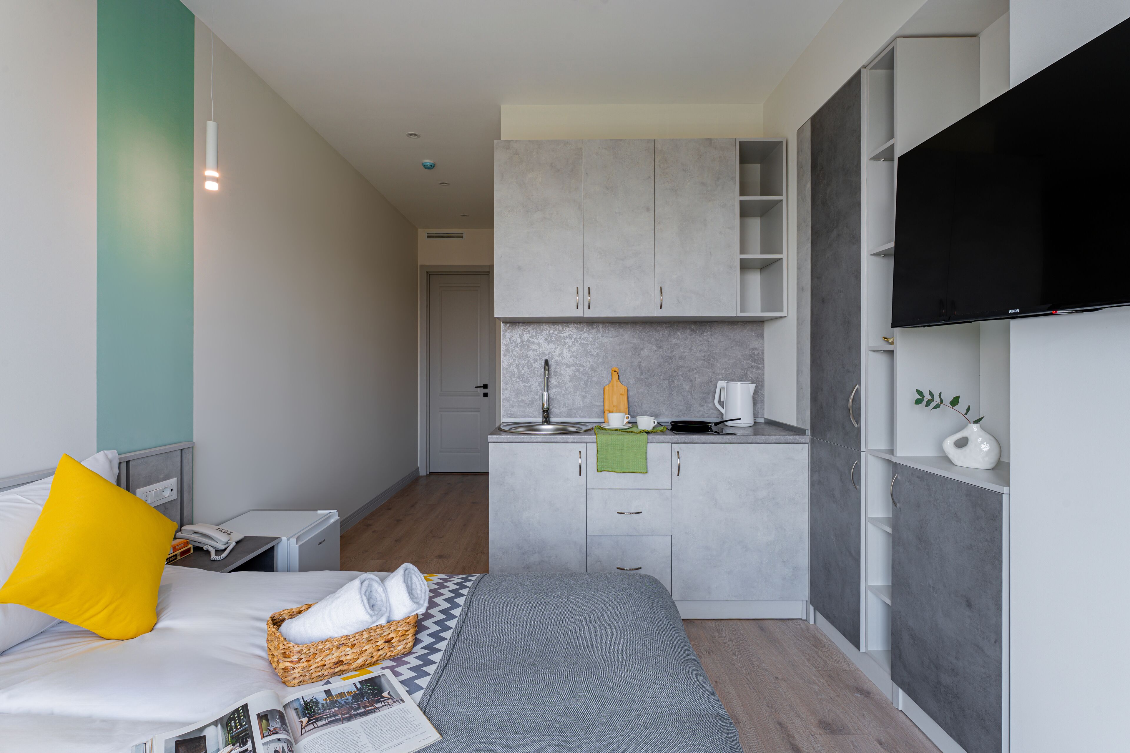 A compact hotel room with a bed, a small kitchenette, a flat-screen TV, and a bathroom.