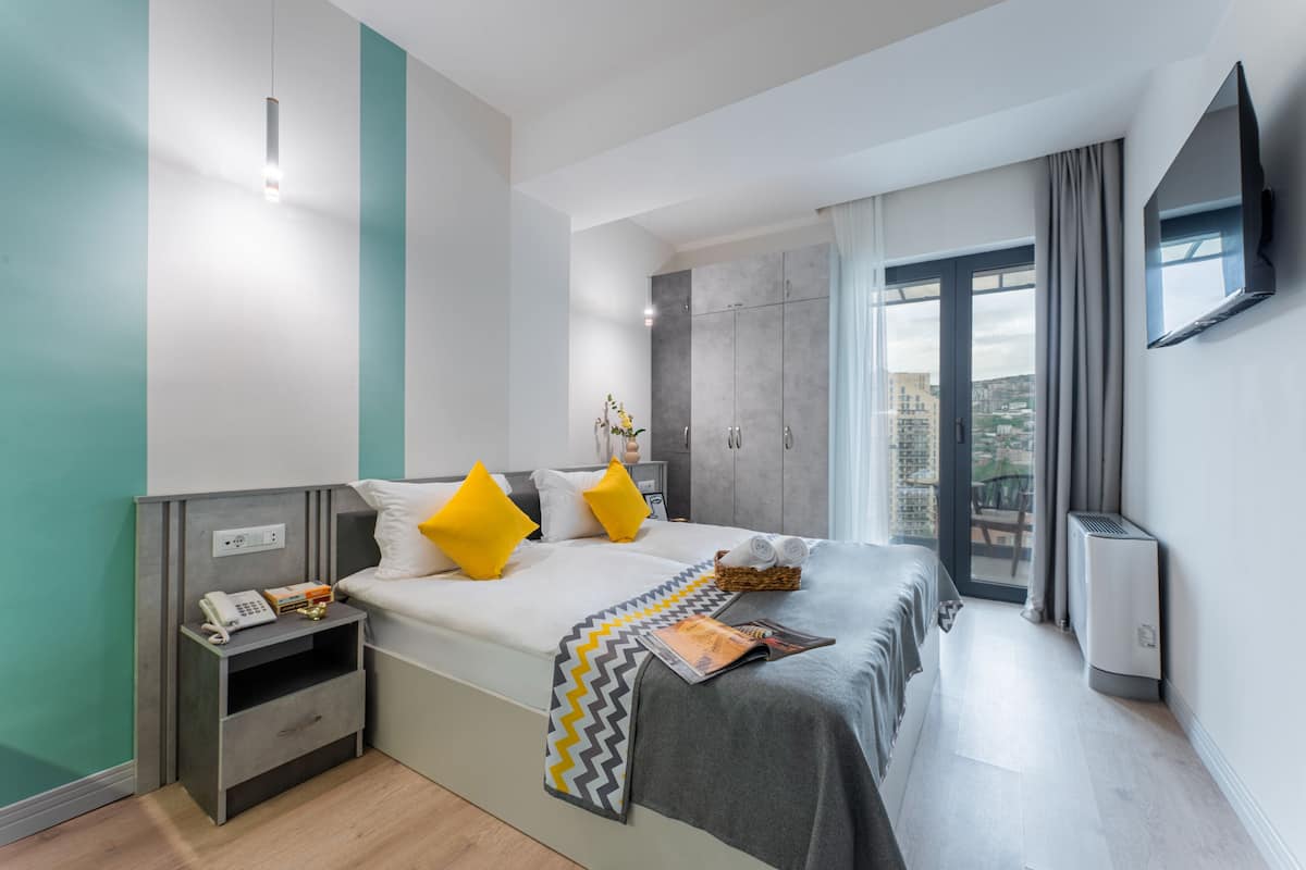 Una habitación de hotel moderna con cama, mesita de noche y vistas de la ciudad a través de un ventanal amplio.