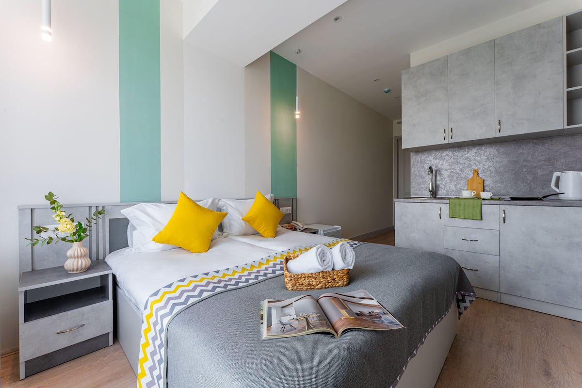 Un hotel moderno con una cama bien hecha, una mesita de noche con lámpara, una pequeña mesa auxiliar y vistas al área de la cocina.