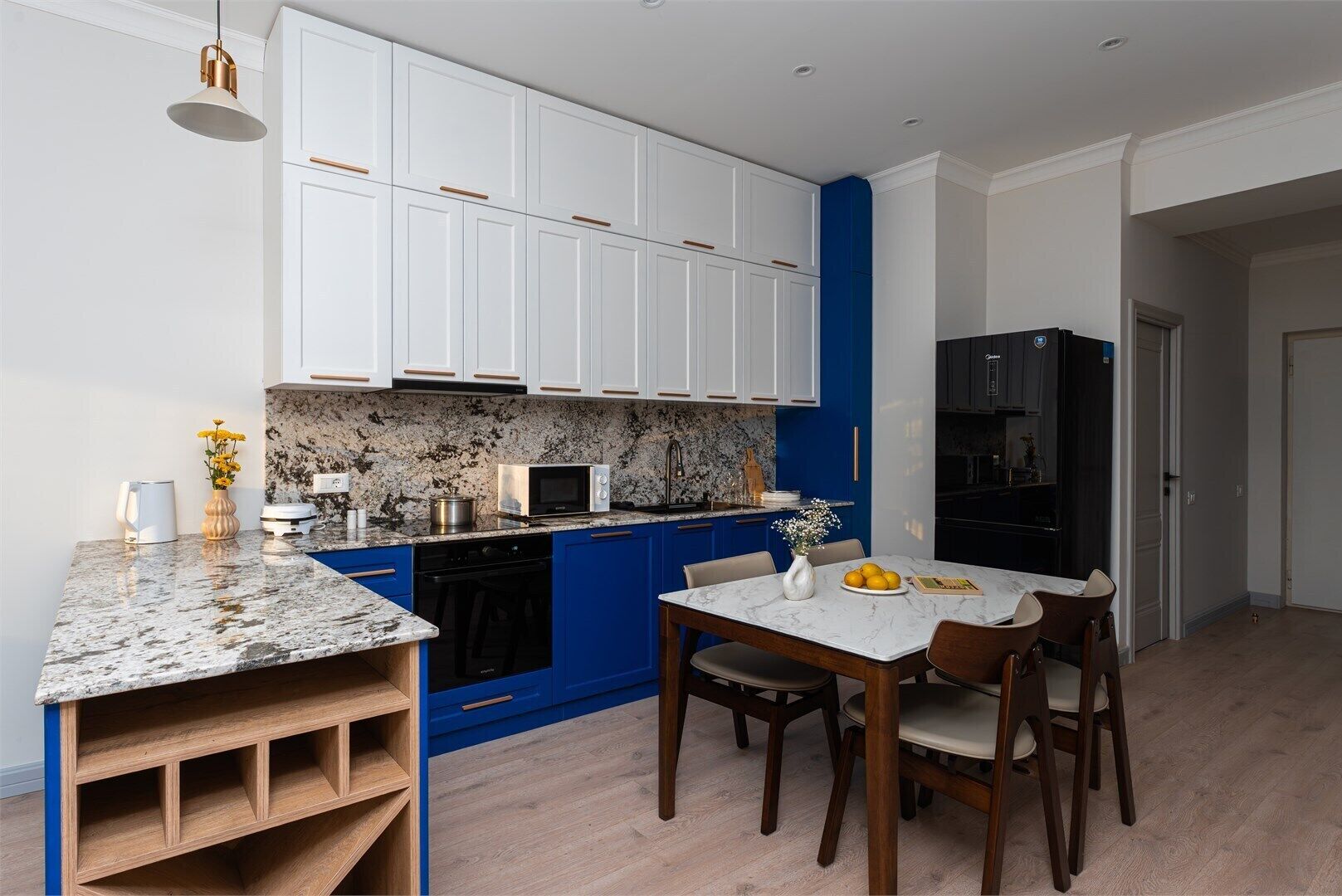 A compact kitchen with white cabinets, a blue accent wall, a marble countertop, and a dining area with a white table and chairs.