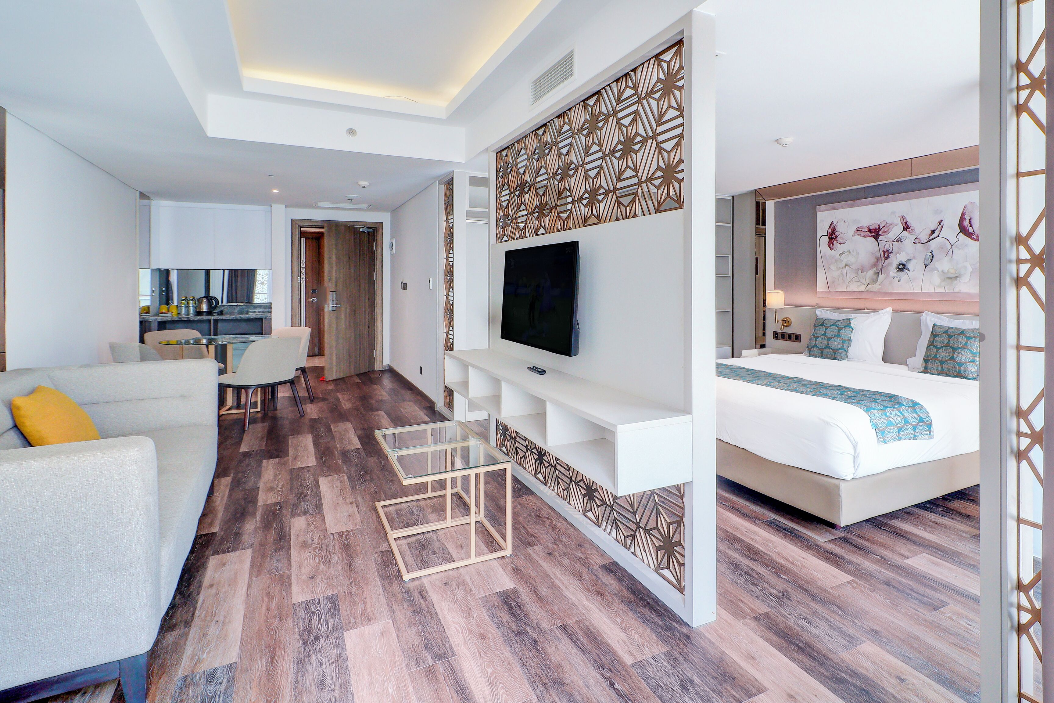 Une chambre d’hôtel moderne avec un grand lit, une télévision, un coin salon et une cuisine à l’arrière-plan.