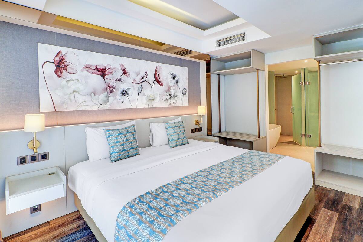 Una camera d'albergo moderna con un letto grande, un comodino, una lampada a parete, un bagno con porta in vetro e un'ampia opera d'arte floreale sulla parete.