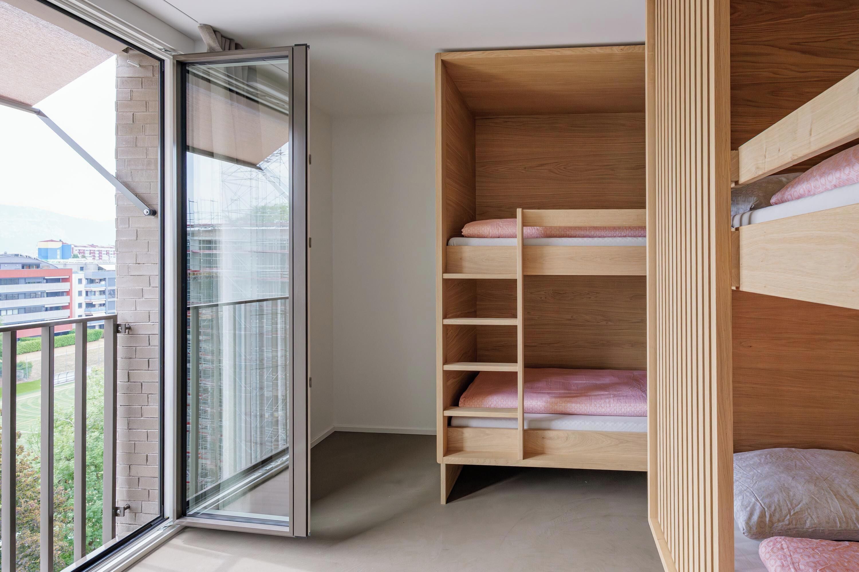A room with a large window, a wooden bunk bed, and a view of buildings outside.