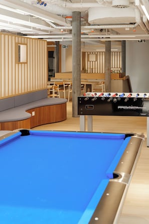 Une table de billard recouverte d’un drap bleu, un coin salon avec un banc incurvé et, au fond, une table de baby-foot.