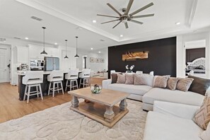 A spacious living area with a large sofa, a coffee table, and a dining area with chairs and a kitchen in the background.