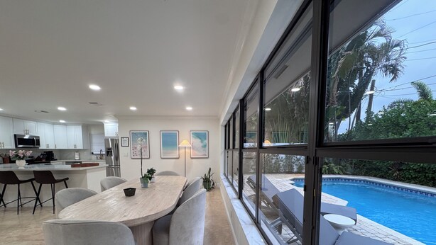 Eine moderne Küche mit weißen Schränken, eine Essecke mit einem Marmortisch und ein großes Fenster mit Blick auf einen Swimmingpool.