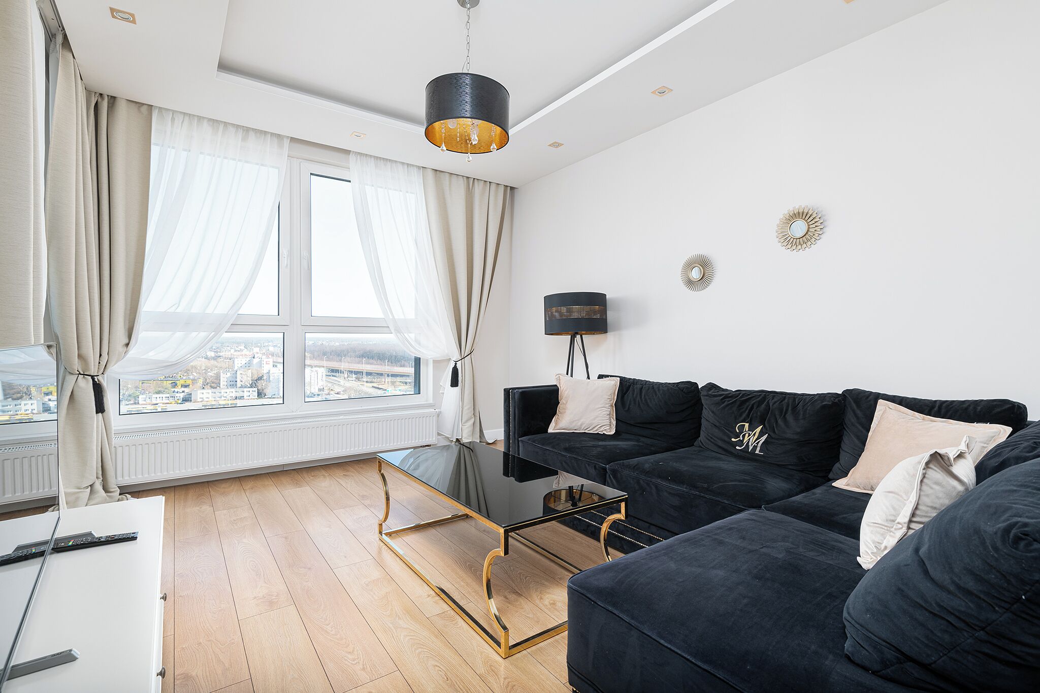 A modern living room with a black sofa, a glass coffee table, and a view of the city through large windows.
