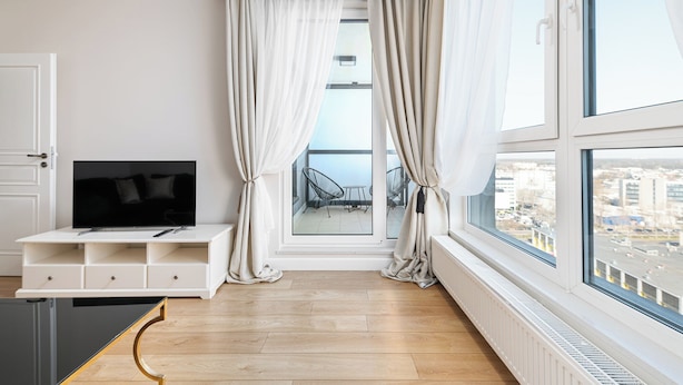 Un salon moderne avec une grande télévision posée sur un meuble blanc, un balcon avec des chaises et une vue sur la ville.
