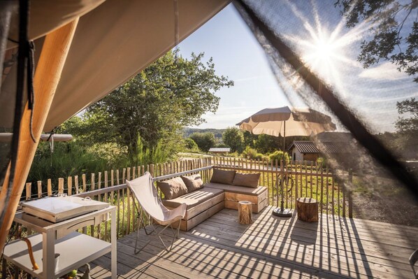 A wooden deck with a sofa, chairs, and a table, overlooking a garden with trees and a distant mountain. - Huttopia Pays de Condrieu  (Tupin-et-Semons)