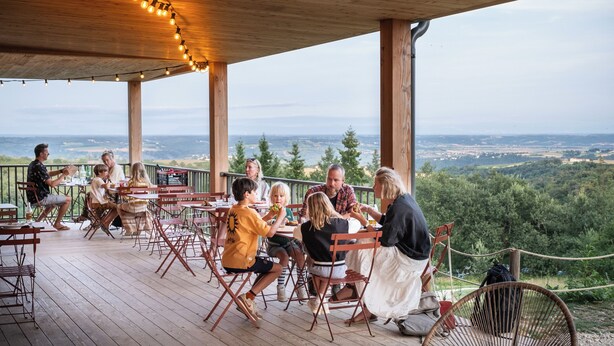 Una familia cenando en una terraza de madera con vistas a un paisaje boscoso.