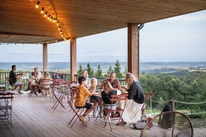 Une famille est assise à une table sur une terrasse en bois, d’où elle peut admirer un paysage boisé.