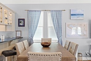 Un coin repas avec une table en bois, des chaises recouvertes d’un tissu beige, un centre de table en forme de bol en bois, et une vue par une fenêtre ornée de rideaux à motifs.