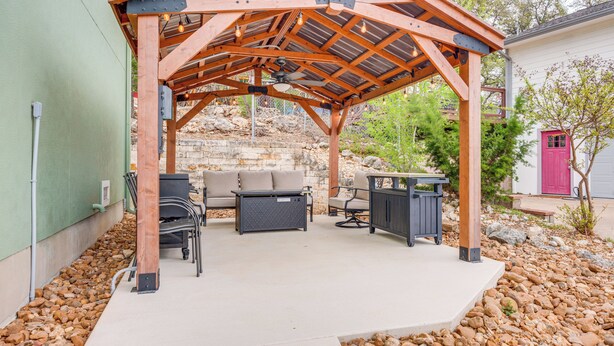 Pátio externo com pérgola de madeira, área de estar e um muro de pedra ao fundo.
