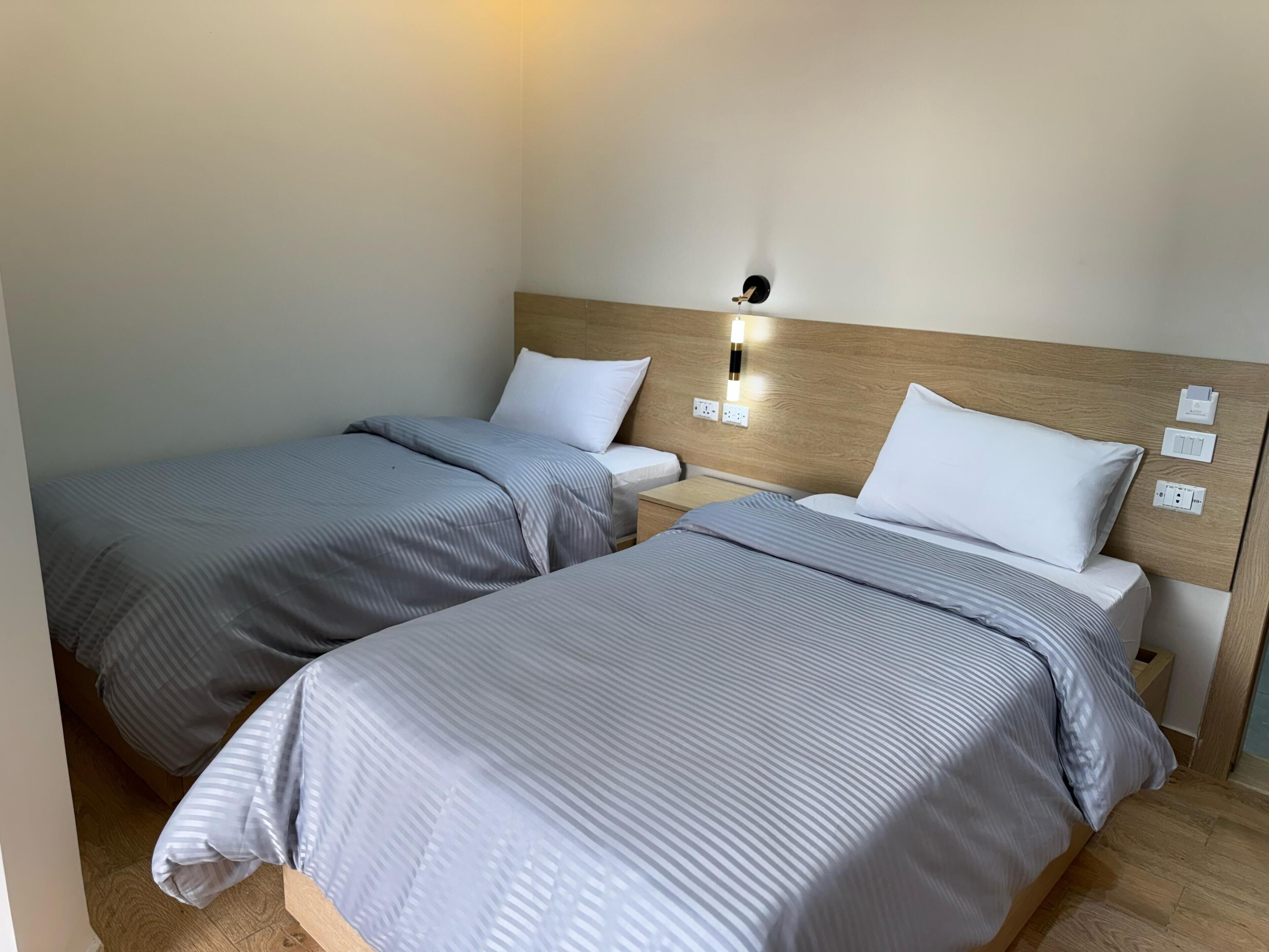 Habitación de hotel con dos camas individuales, cada una con cabecera de madera y mesita de noche. Hay un aplique en la pared sobre las camas.