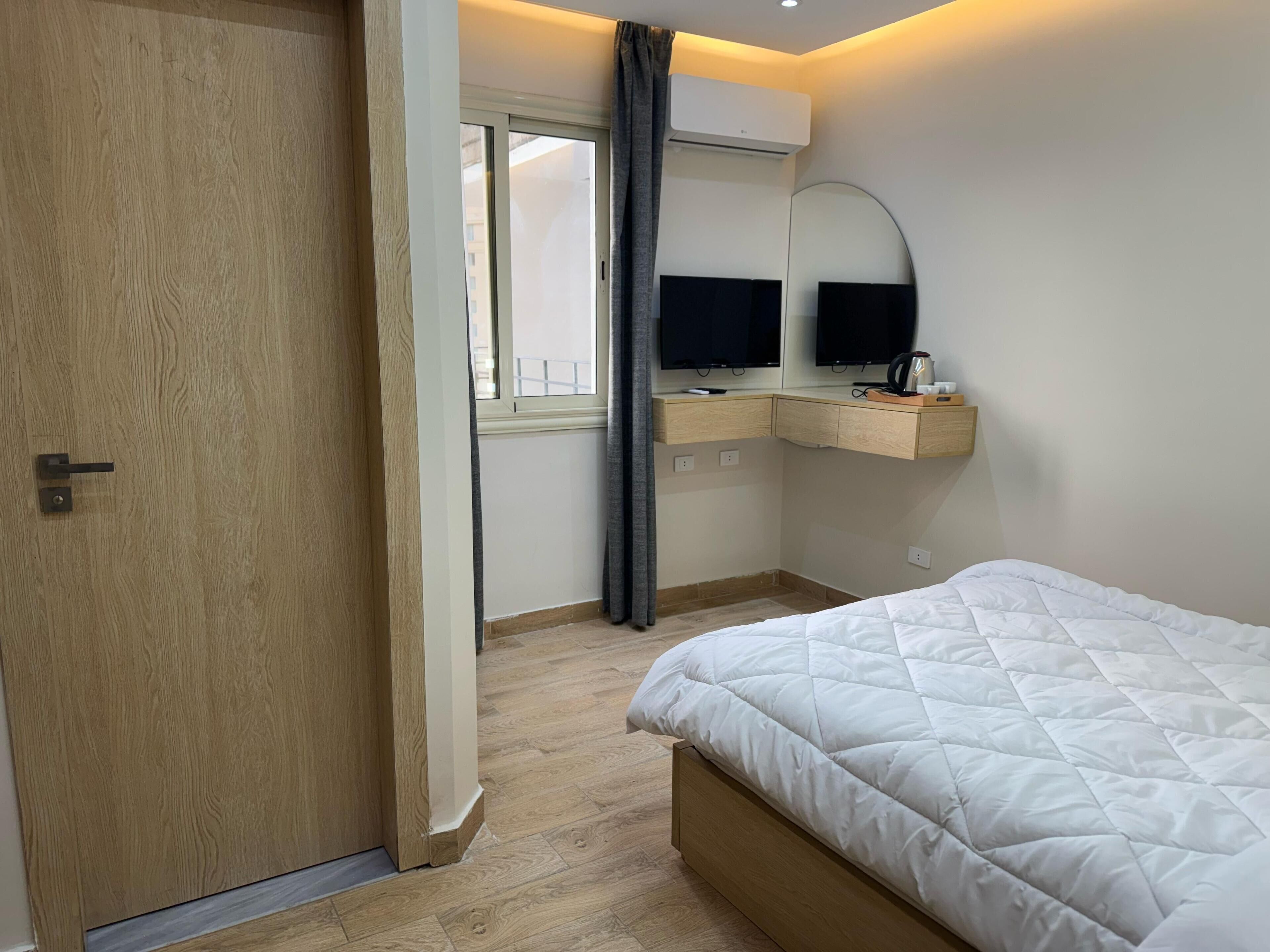 Un dormitorio con puerta de madera, cama con edredón acolchado blanco, escritorio con televisor de pantalla plana, espejo y ventana con cortinas.