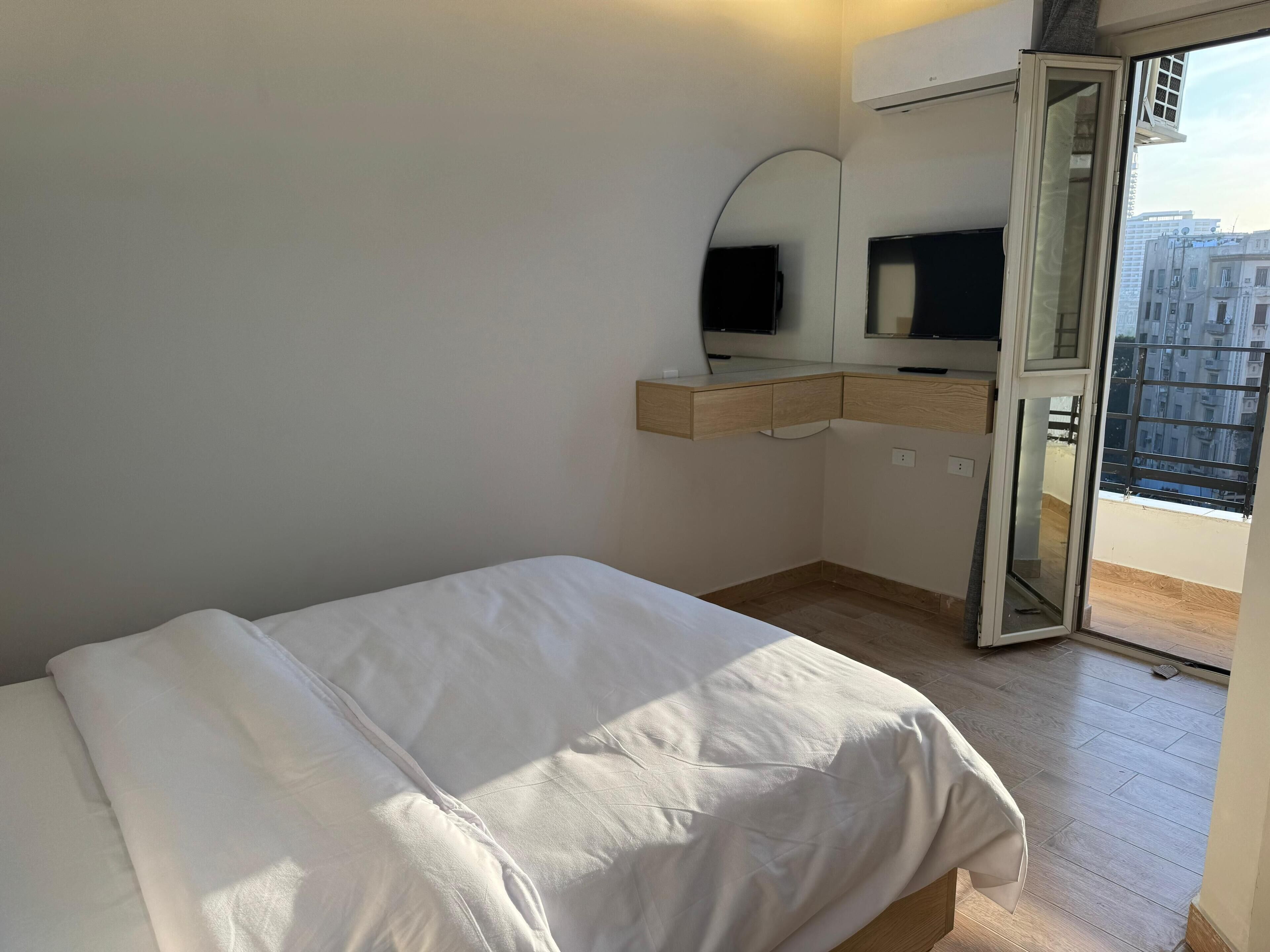 Un dormitorio con cama, un escritorio de madera, un espejo y una puerta corrediza de vidrio a un balcón.