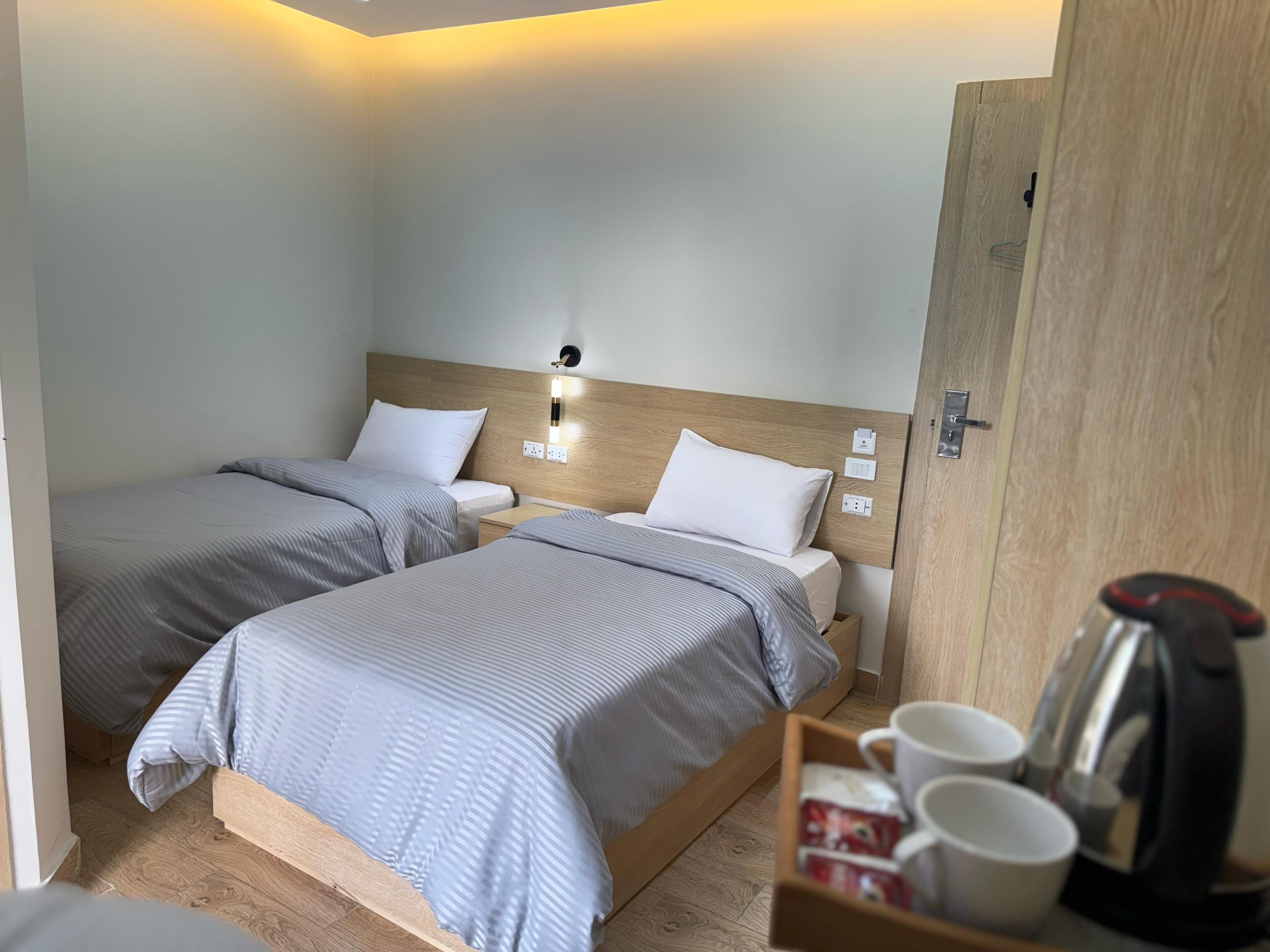 A hotel room with two single beds, a wooden headboard, bedside lamps, a coffee maker, and a tray with cups and a kettle.