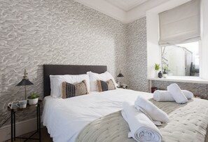 Quarto com parede texturizada, cama com roupa de cama branca, abajur, janela com vista e mesa de cabeceira com uma planta.