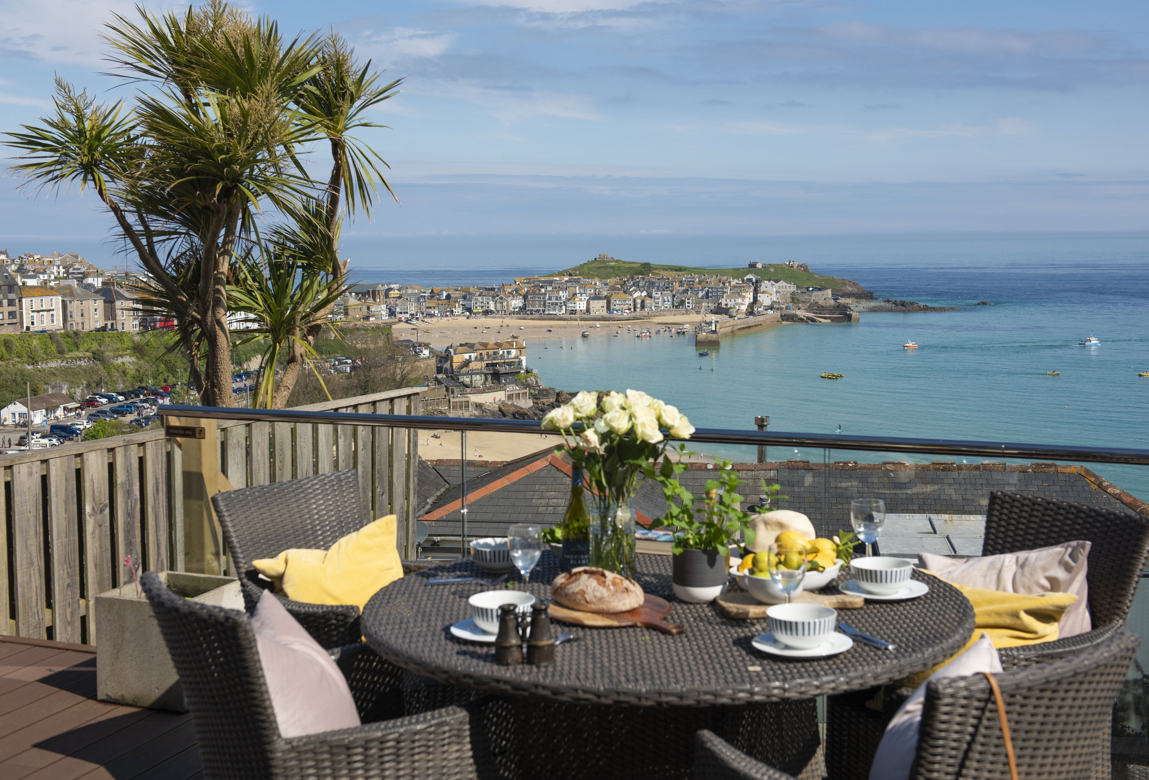 A dining table with chairs, a vase of flowers, and a view of the sea and town.