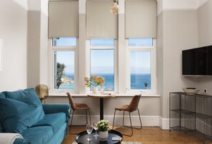 Uma sala de estar com sofá, uma mesa de jantar com cadeiras, televisão e vista para o mar.