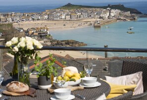 Vista al mar desde un balcón con mesa lista para comer, con vista a una playa y un pueblo histórico.