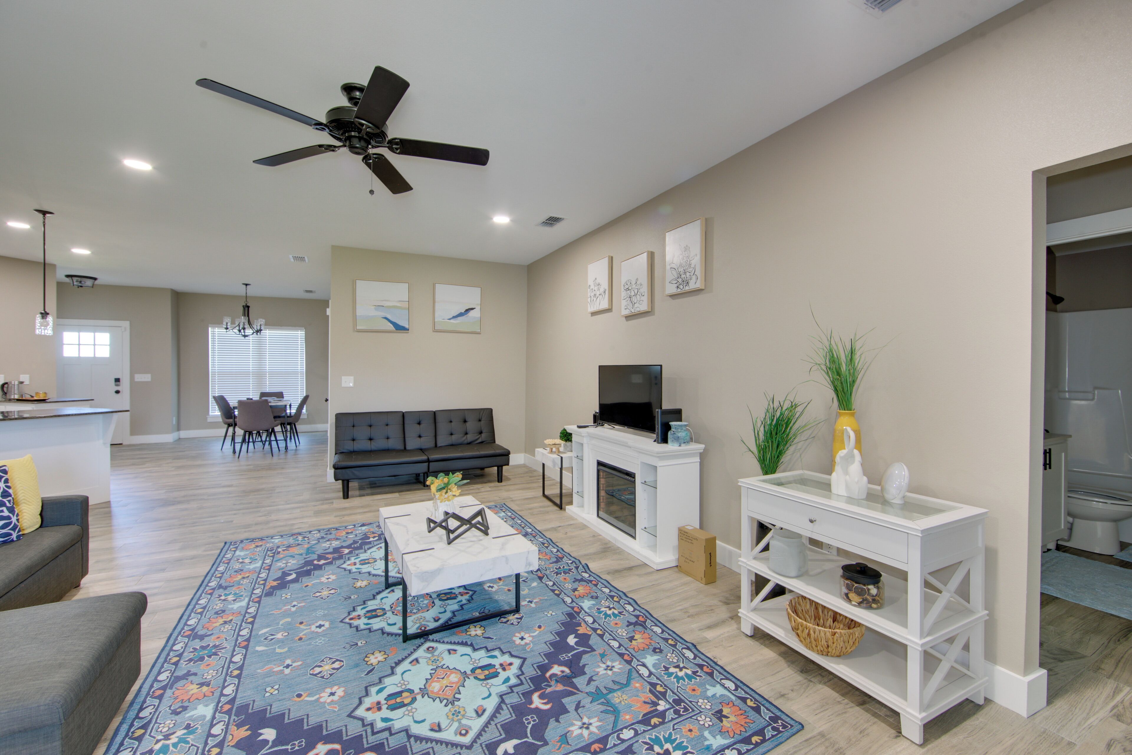 A modern living room with a sofa, coffee table, and TV. There is a fireplace, a ceiling fan, and a dining area in the background.