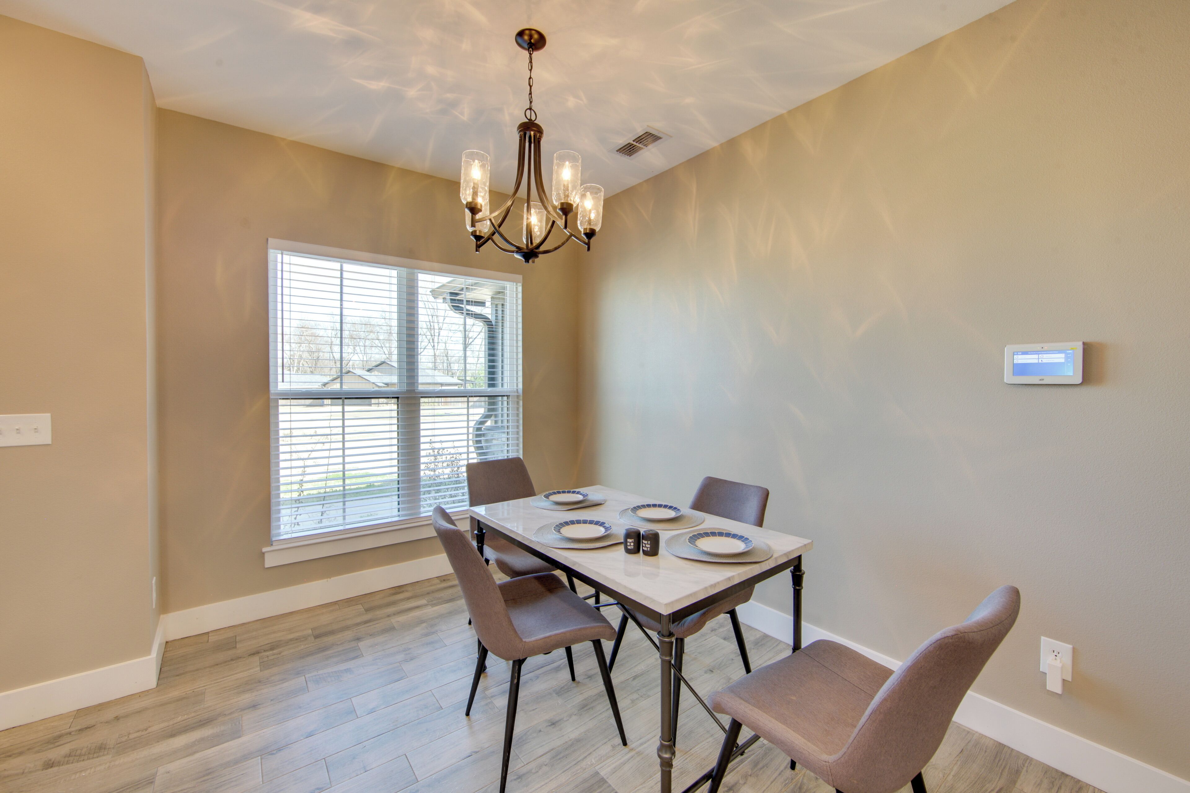 A dining area with a table set for four, surrounded by chairs, a chandelier, and a window with blinds.
