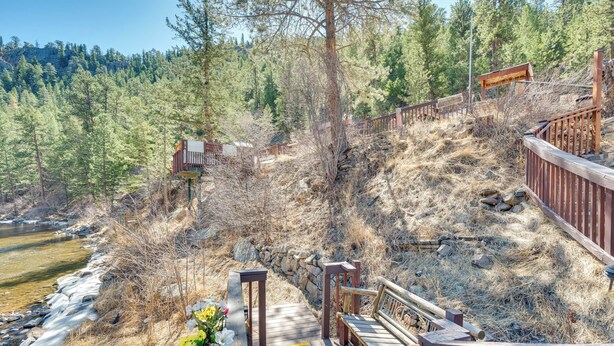A wooden deck with a stone wall, overlooking a river and forested area. - Fish, Hike & Unwind: Drake Cabin w/ Hot Tub! (Drake)