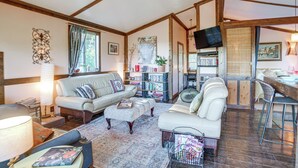 Un salon chaleureux avec un canapé, un pouf et une table basse. On y trouve une bibliothèque remplie de livres et d’objets décoratifs, un téléviseur fixé au mur, ainsi qu’un coin repas avec une table et des chaises.