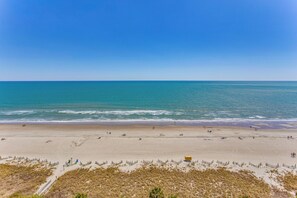 A wide sandy beach with gentle waves, a clear sky, and sparse vegetation in the foreground. - Sun, Sand & Sea Views: Myrtle Beach Condo (Myrtle Beach)