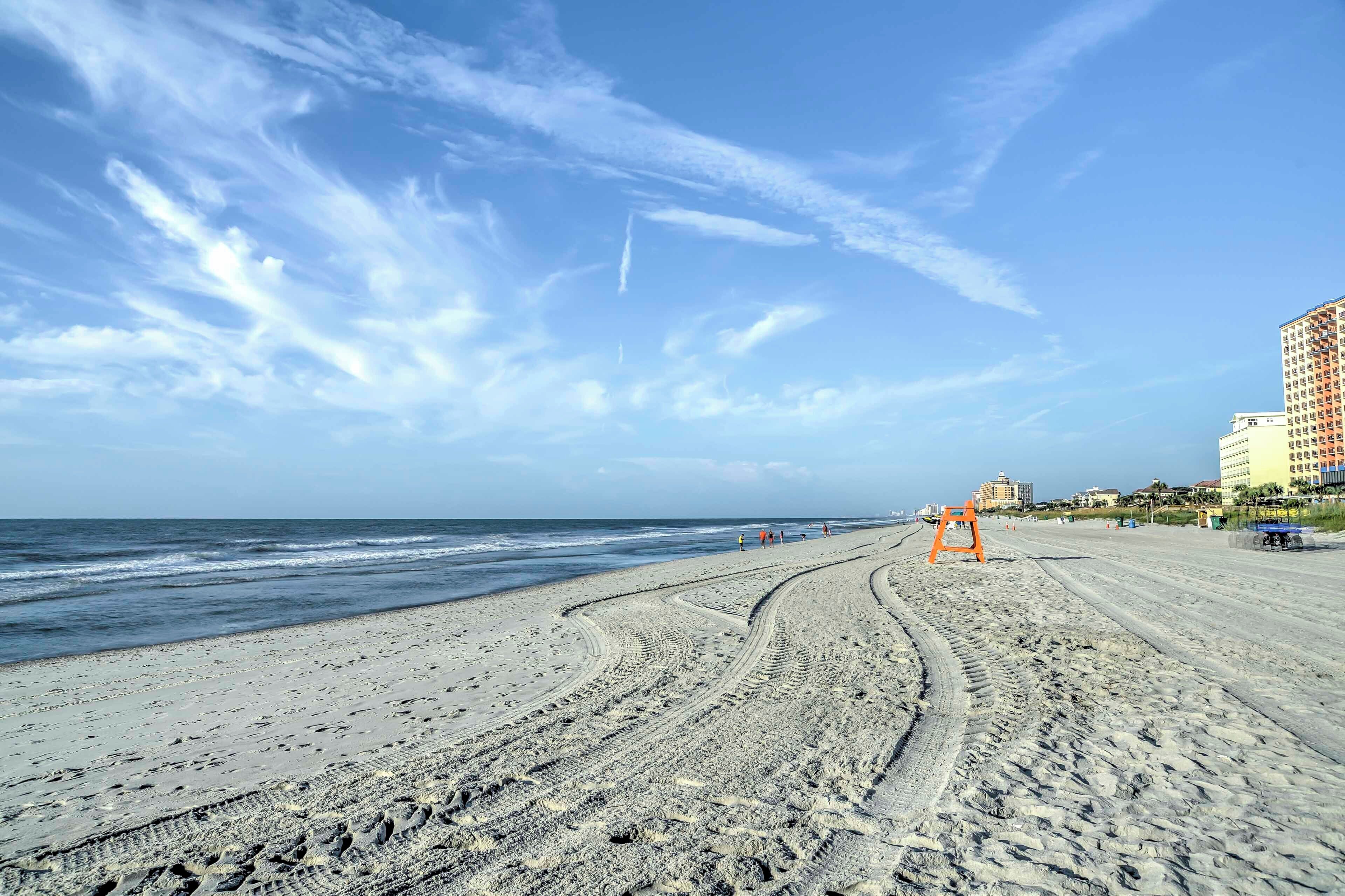 Una playa con marcas de neumáticos, una torre de salvavidas y edificios al fondo.