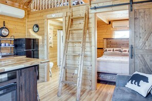 Interieur van een houten hutje met een keukenhoek, een ladder, een bed met een beer-kussen en een houten deur.