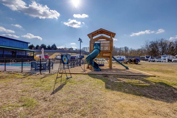 A playground with a slide and a wooden structure, surrounded by a fenced area with a pool and a building in the background.