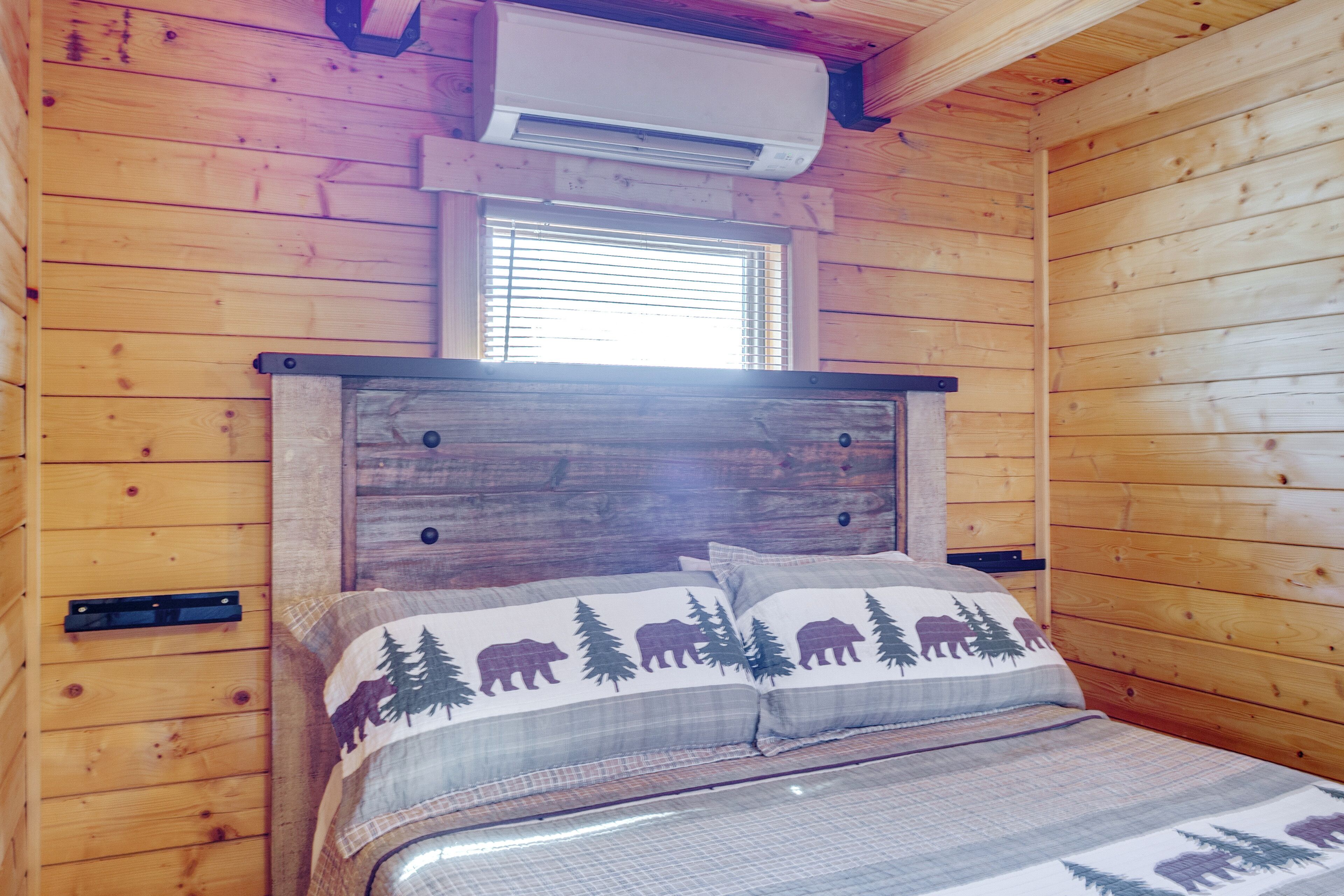 A wooden cabin bedroom with a bed, a window with blinds, and an air conditioning unit.
