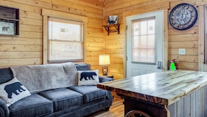 A cozy cabin interior with a wooden ceiling, a leather sofa with bear-themed pillows, a wooden table, and a wall clock. - Fire Pit + Pool Access: Cozy Cabin in Sevierville! (Sevierville)