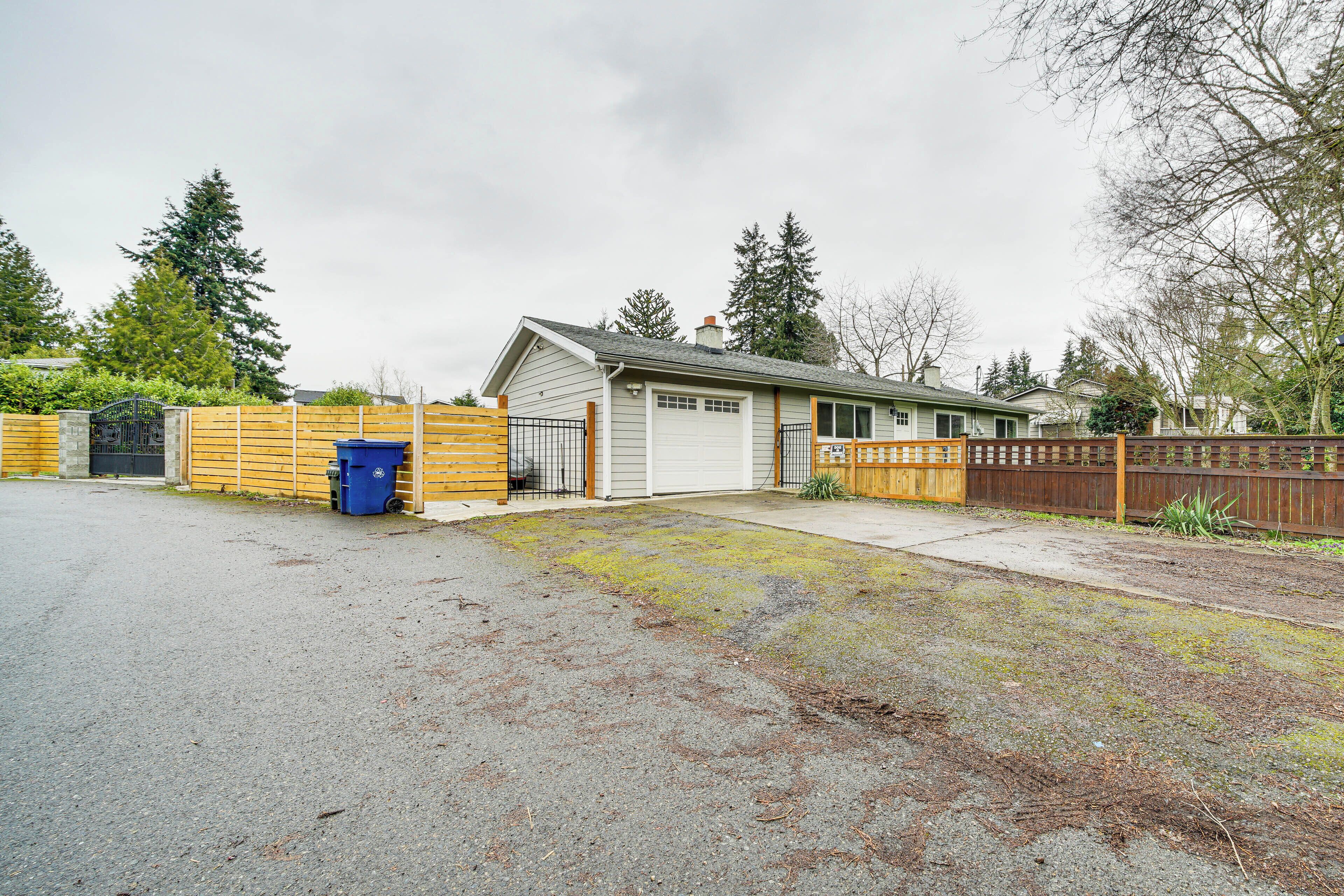 A residential garage with a wooden fence, a blue trash bin, and a driveway leading to a house.