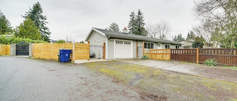 A residential garage with a wooden fence, a blue trash bin, and a driveway leading to a house.