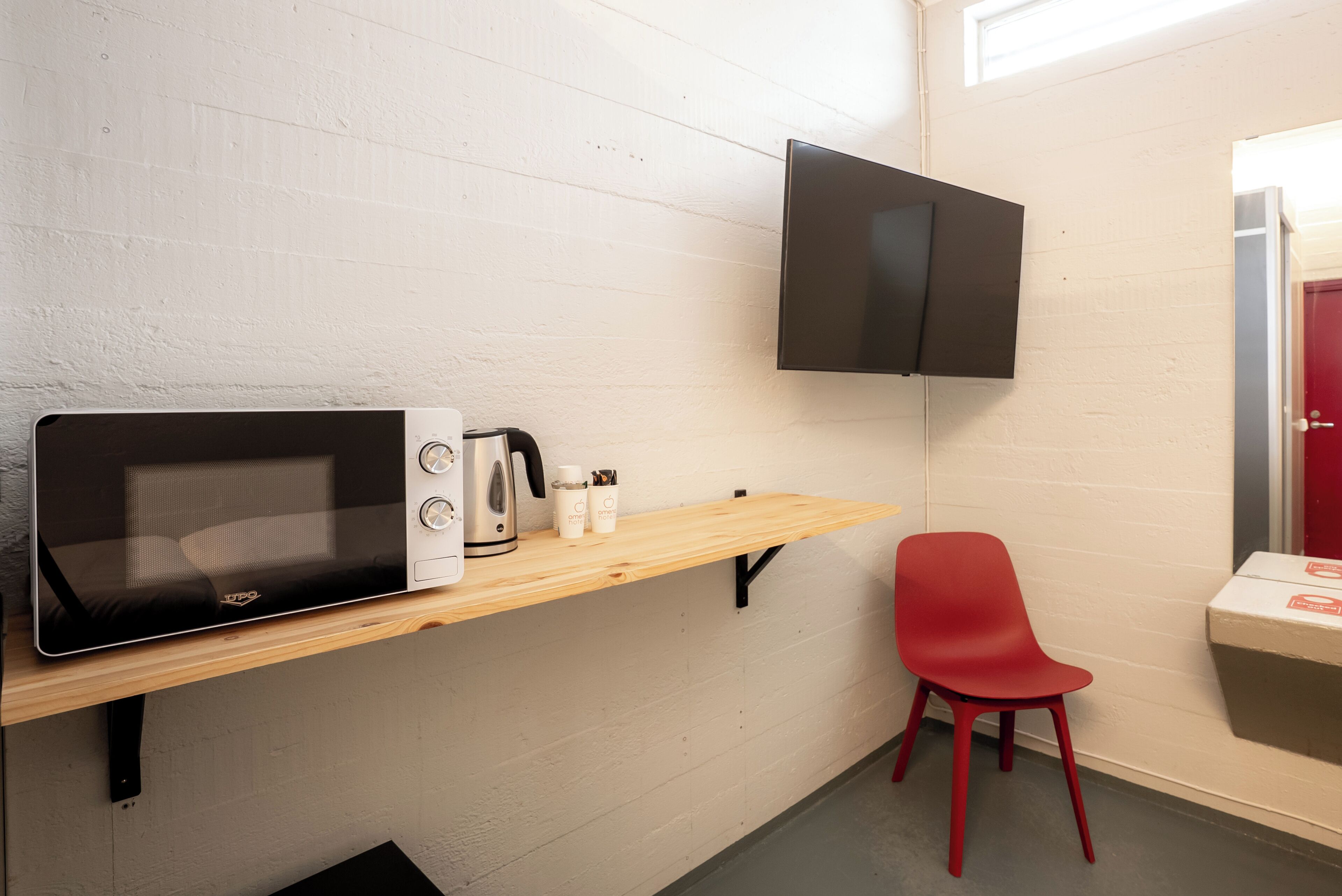 A kitchenette with a microwave, kettle, and coffee maker on a wooden shelf, a red chair, and a flat-screen TV mounted on the wall.